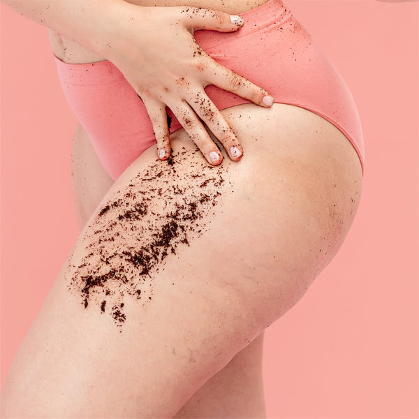 A hand pressing the coffee scrub from the frank body Baby Mama Kit over a hip and thigh above pink underwear on a pink background.