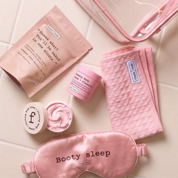 Flat lay of the frank body Baby Mama Kit with the coffee scrub sachet, open firming body cream, rosehip scrub, waffle towel and Booty sleep eye mask on tiles.