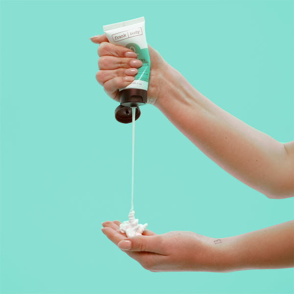 A hand squeezes a tube of body wash, dispensing white foam into the other hand against a solid light aqua background. The tube has a white and green label with the brand name 'Frank body' visible.