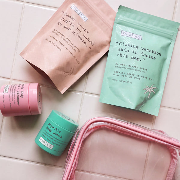 Flat lay of the frank body Scrub Stars Kit on white tiles with the Original and Coconut Coffee Scrub pouches, two scrub tubs and a clear pink travel bag.