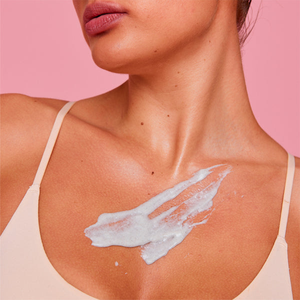 White scrub from the frank body Scrub Stars Kit smeared across a woman's chest and collarbone as she stands in a cream bra on a pink background.