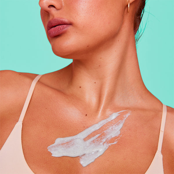 Close-up of a woman's neck and upper chest with a white cream or sunscreen swipe applied on her skin, wearing a light beige bra against a solid mint green background.