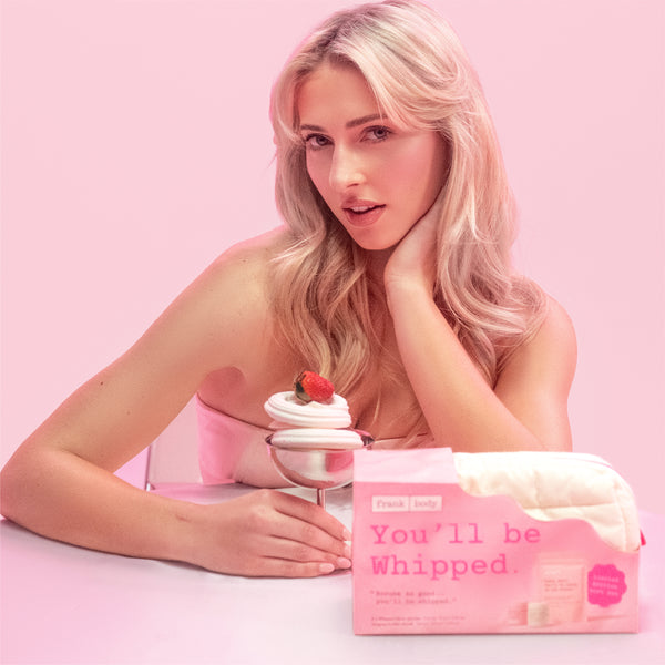 A blonde woman in a pink top leans over a table with a pink product box in front of her. The box reads 'Frank | Body You'll be Whipped.