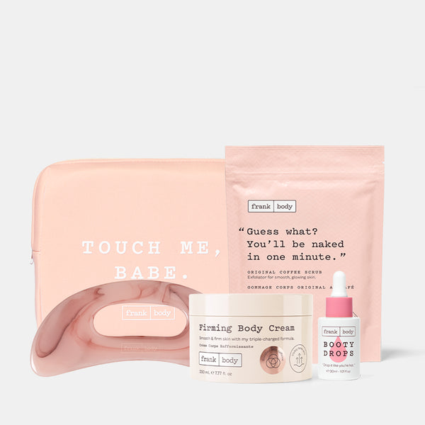A Frank Body self-care set featuring a blush pink pouch with 'TOUCH ME, BABE.' text, a matching pink curved tray, a cream jar labeled 'Firming Body Cream,' a small white bottle labeled 'BOOTY DROPS,' and a pink stand-up pouch with a quote, arranged on a light gray background.