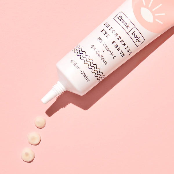 A tube of frank body Brightening Eye Serum with drops of the serum beneath it on a pink background.